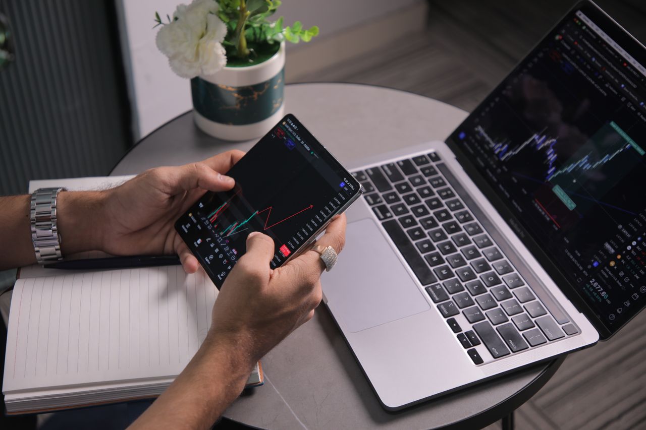 Un homme examine les tendances boursières sur un ordinateur portable et une application de trading mobile dans un espace de travail moderne.